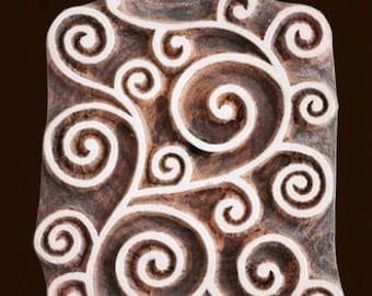 May include: A rectangular wooden block with a carved swirling pattern. The design features interconnected spirals and curls in a light colour against a dark brown background. This block is likely used for printing or stamping.