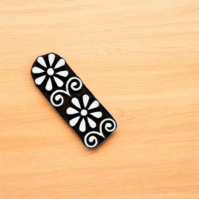 Puede incluir: Un sello de bloque floral en blanco y negro. El sello presenta dos flores blancas en forma de margarita y remolinos decorativos sobre una base negra rectangular. El sello est&aacute; sobre una superficie de madera marr&oacute;n claro.