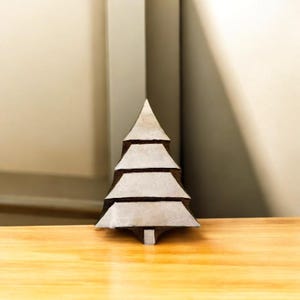 Könnte beinhalten: Ein kleiner, dekorativer Weihnachtsbaum aus geschichtetem, hellgrauem Material. Der Baum hat eine dreieckige Form mit horizontalen Ebenen und steht auf einer Holzoberfläche. Eine festliche Weihnachtsdekoration.