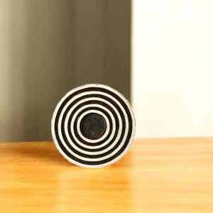 K&ouml;nnte beinhalten: Ein rundes Dekorationsobjekt mit konzentrischen schwarzen und wei&szlig;en Streifen. Die Mitte ist dunkel gef&auml;rbt. Das Objekt liegt auf einer hellen Holzoberfl&auml;che.