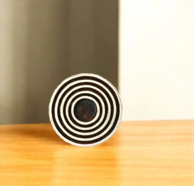 K&ouml;nnte beinhalten: Ein rundes Dekorationsobjekt mit konzentrischen schwarzen und wei&szlig;en Streifen. Die Mitte ist dunkel gef&auml;rbt. Das Objekt liegt auf einer hellen Holzoberfl&auml;che.