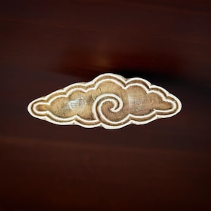 Puede incluir: Un bloque de madera tallado en forma de nube con un diseño de espiral. El bloque es de color marrón claro con líneas blancas que delinean la forma de la nube.