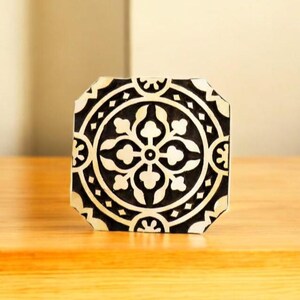 May include: A square, decorative tile with a black and white floral pattern. The design features a central flower motif surrounded by circular and geometric elements. The tile is octagonal in shape and sits on a wooden surface.
