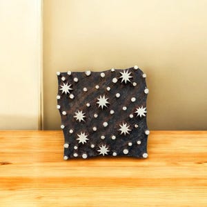 May include: A square, dark brown wooden block with a celestial design. The block features white, star-shaped and dot-shaped patterns. The block is sitting on a wooden surface.