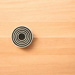 K&ouml;nnte beinhalten: Ein rundes, schwarz-wei&szlig;es dekoratives Objekt mit konzentrischen Kreisen. Das Objekt befindet sich auf einer hellbraunen Holzoberfl&auml;che. Das Design zeigt abwechselnd schwarze und wei&szlig;e B&auml;nder, die ein ziel&auml;hnliches Muster erzeugen.