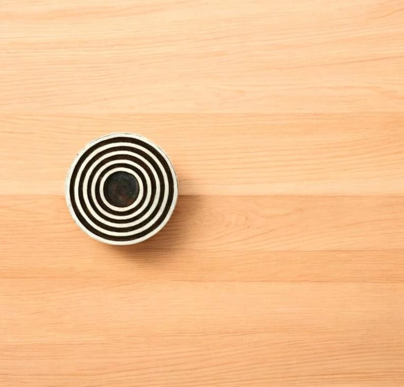 K&ouml;nnte beinhalten: Ein rundes, schwarz-wei&szlig;es dekoratives Objekt mit konzentrischen Kreisen. Das Objekt befindet sich auf einer hellbraunen Holzoberfl&auml;che. Das Design zeigt abwechselnd schwarze und wei&szlig;e B&auml;nder, die ein ziel&auml;hnliches Muster erzeugen.