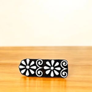 Puede incluir: Un bloque de madera negro rectangular con un dise&ntilde;o floral. El bloque presenta dos flores blancas en forma de margarita y volutas blancas. El bloque est&aacute; sobre una superficie de madera.