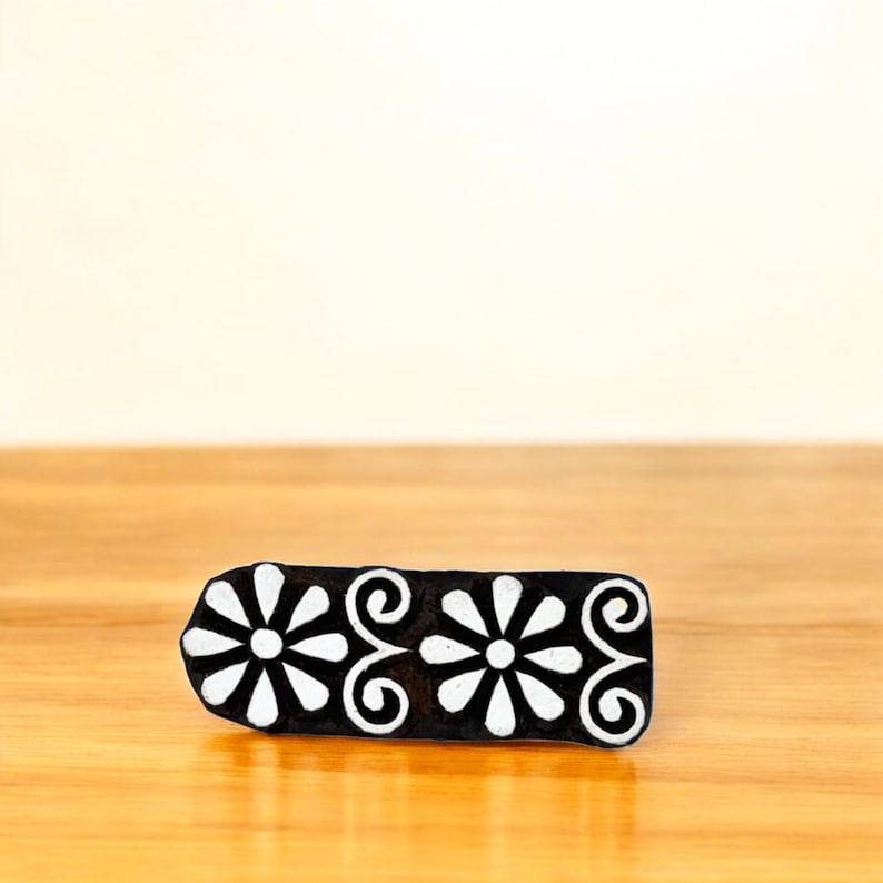 Puede incluir: Un bloque de madera negro rectangular con un dise&ntilde;o floral. El bloque presenta dos flores blancas en forma de margarita y volutas blancas. El bloque est&aacute; sobre una superficie de madera.
