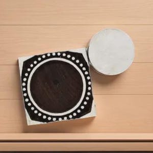 Può includere: Una piastrella decorativa quadrata in bianco e nero con un design circolare e un bordo a pois, accanto a una piastrella rotonda bianca. Le piastrelle sono su una superficie di legno marrone chiaro.