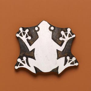 May include: A white frog silhouette carved into a wooden block. The block is painted black and has a rough, textured surface.