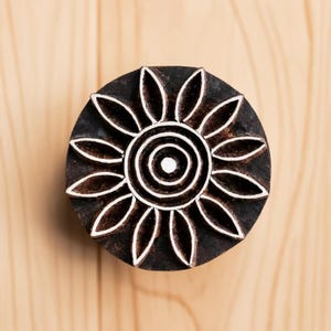 May include: A round wooden block with a carved design of a sun with a circle in the center. The sun has 12 rays and the circle has three concentric circles.