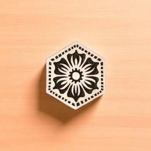 May include: A hexagonal coaster with a black floral design. The coaster has a white border with dotted accents. The floral pattern features a central circle surrounded by stylized petals. The coaster is placed on a light orange wooden surface.