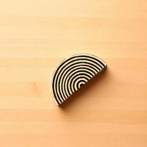 Op de afbeelding: Een halfrond decoratief object met een zwart-wit gestreept patroon. De strepen zijn concentrisch, waardoor een visueel beeld van een halve cirkel regenboog ontstaat. Het object staat tegen een lichte houtnerf achtergrond.