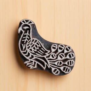 Puede incluir: Un bloque de madera tallada con un diseño de pavo real. El bloque es de color gris oscuro con un diseño de pavo real blanco. El pavo real tiene una cola y un cuerpo detallados. El bloque está sobre una superficie de madera marrón claro.
