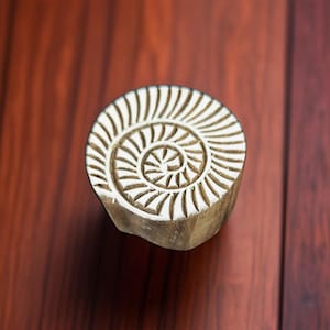 May include: A white wooden block with a carved spiral design. The block is used for printing patterns on fabric.