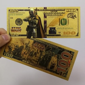 May include: Two gold-colored paper bills with Star Wars characters. The first bill features Boba Fett and the text "FEDERAL RESERVE NOTE GOLD 999999 STAR WARS 100". The second bill features a group of Star Wars characters and the text "UNITED STATES OF AMERICA STAR WARS 100".