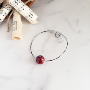 May include: A silver wire bracelet featuring a red and black marbled bead. The bracelet has a spiral design at the top. The background is white marble. Rolled sheet music is visible in the background.