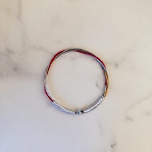 Può includere: Un bracciale circolare realizzato con un filo metallico argentato avvolto. Il design incorpora un filo rosso vibrante intrecciato attraverso le bobine metalliche, con due distinti segmenti argentati a forma di molla posizionati alla chiusura.