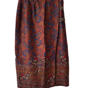 May include: A vintage wrap skirt with a paisley pattern in shades of red, blue, and gold. The skirt features a brown border with a hunting scene and brown leather buckle closures. The skirt is hanging on a white hanger.