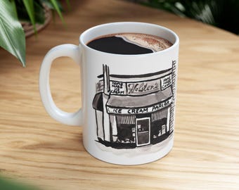 May include: White ceramic mug with a black and white illustration of an ice cream parlor. The mug is filled with dark coffee. The ice cream parlor has a sign that says "Holsten's Ice Cream Parlor" and "Home Made Ice Cream".