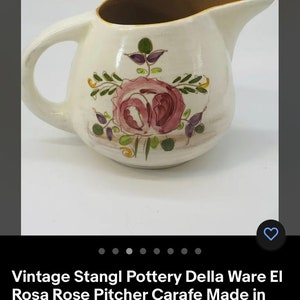 Puede incluir: Una jarra de cerámica blanca con un borde marrón y un diseño floral con rosas rosas, hojas verdes y detalles morados. La jarra es una jarra vintage Stangl Pottery Della Ware El Rosa Rose hecha en los Estados Unidos.