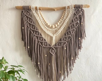 Puede incluir: Un tapiz de macramé de pared con un esquema de color marrón y blanco. El tapiz presenta un diseño geométrico con cuentas de madera y flecos.