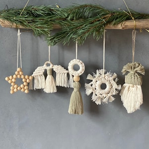 May include: A set of six handmade macrame ornaments hanging from a driftwood branch. The ornaments include a star, two angels, a circle, a snowflake, and a gnome. The ornaments are made with natural fibres and have a rustic, bohemian style.