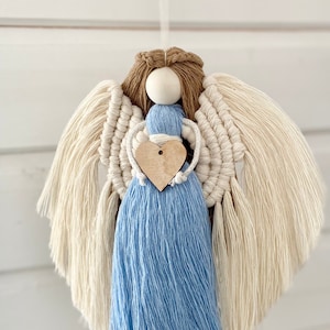 May include: A handcrafted macramé angel featuring cream-coloured wings, a light blue dress, and a wooden heart. The angel has brown hair and a wooden bead head. The wings are made of knotted cotton cord, and the dress is a tassel of blue yarn.