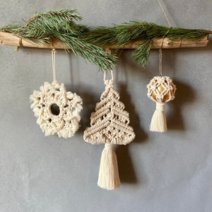 May include: A rustic Christmas decoration featuring a wooden branch adorned with green pine needles. Three handmade macrame ornaments hang below: a snowflake, a Christmas tree, and a sphere, all in natural off-white tones.