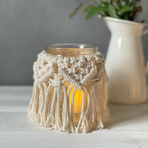 以下が含まれることがあります： ガラス製のキャンドルホルダーに、かぎ針編みで作ったカバーとフリンジが付いています。キャンドルホルダーには灯りがともったキャンドルが入っており、白い木製の表面の上に置かれています。