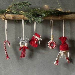Puede incluir: Adornos navideños hechos a mano colgados de una rama de madera con vegetación. Los adornos incluyen un bastón de caramelo, un reno, un calcetín, una corona y un gnomo, todos en rojo, blanco y gris. Cuelgan de cuerdas blancas.