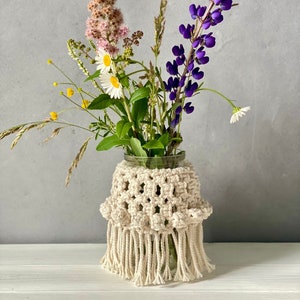 Peut inclure: Un vase en verre avec un couvre-vase en crochet de couleur crème. Le vase est rempli d'un bouquet de fleurs sauvages, dont des lupins violets, des spirées roses et des marguerites blanches.