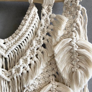 Puede incluir: Un tapiz de macram&eacute; blanco con una barra de madera natural en la parte superior. El macram&eacute; presenta una variedad de nudos y texturas, incluyendo flecos y borlas.