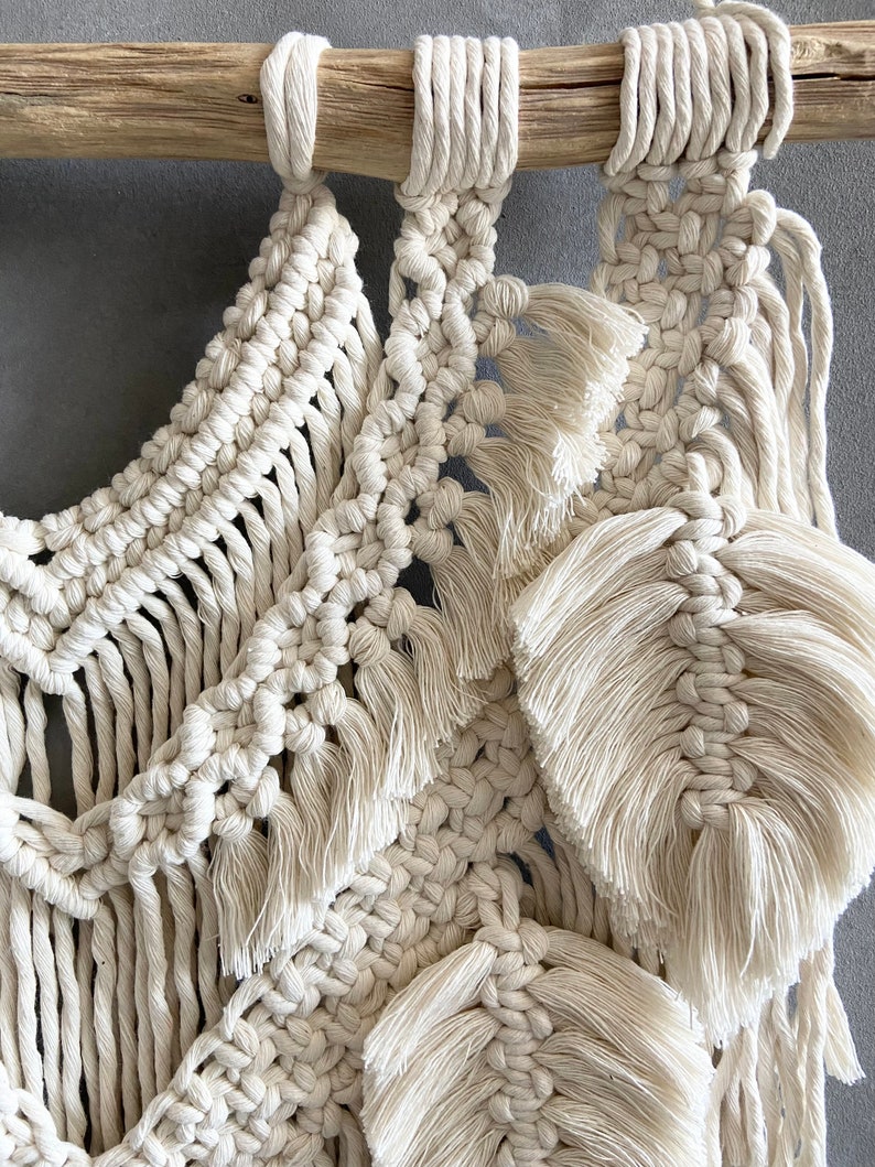 Puede incluir: Un tapiz de macram&eacute; blanco con una barra de madera natural en la parte superior. El macram&eacute; presenta una variedad de nudos y texturas, incluyendo flecos y borlas.
