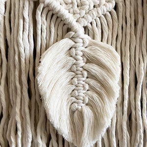 Puede incluir: Primer plano de un tapiz de macram&eacute; con un dise&ntilde;o de pluma. El tapiz est&aacute; hecho con cord&oacute;n de algod&oacute;n de color natural y tiene un detalle de flecos.