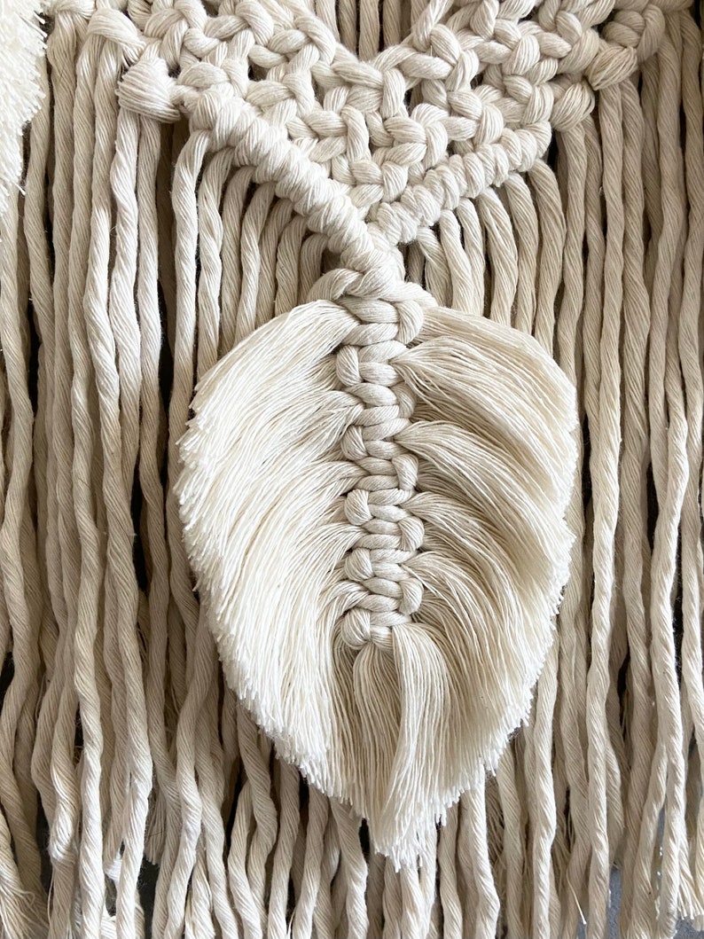 Puede incluir: Primer plano de un tapiz de macram&eacute; con un dise&ntilde;o de pluma. El tapiz est&aacute; hecho con cord&oacute;n de algod&oacute;n de color natural y tiene un detalle de flecos.