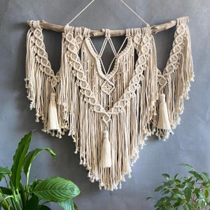以下が含まれることがあります： 幾何学模様の大きな白いマクラメの壁掛け。上部に木製の棒があり、綿のコードで作られています。