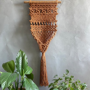 Könnte beinhalten: Ein brauner Makramee-Wandbehang mit einem Holzstab oben und einem langen Quasten unten. Der Behang ist aus einem dicken Seil gefertigt und hat ein geometrisches Muster.