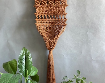 May include: A brown macrame wall hanging with a wooden dowel at the top and a long tassel at the bottom. The hanging is made with a thick cord and features a geometric pattern.