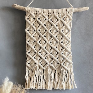 May include: A macrame wall hanging with a diamond pattern and fringe. The hanging is made of natural cotton cord and is attached to a piece of driftwood.