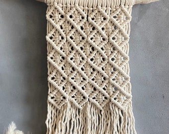 May include: A macrame wall hanging with a diamond pattern and fringe. The hanging is made of natural cotton cord and is attached to a piece of driftwood.