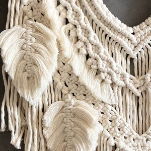 Puede incluir: Un tapiz de macram&eacute; blanco con un dise&ntilde;o geom&eacute;trico y detalles de flecos. El tapiz presenta una serie de hojas tejidas.