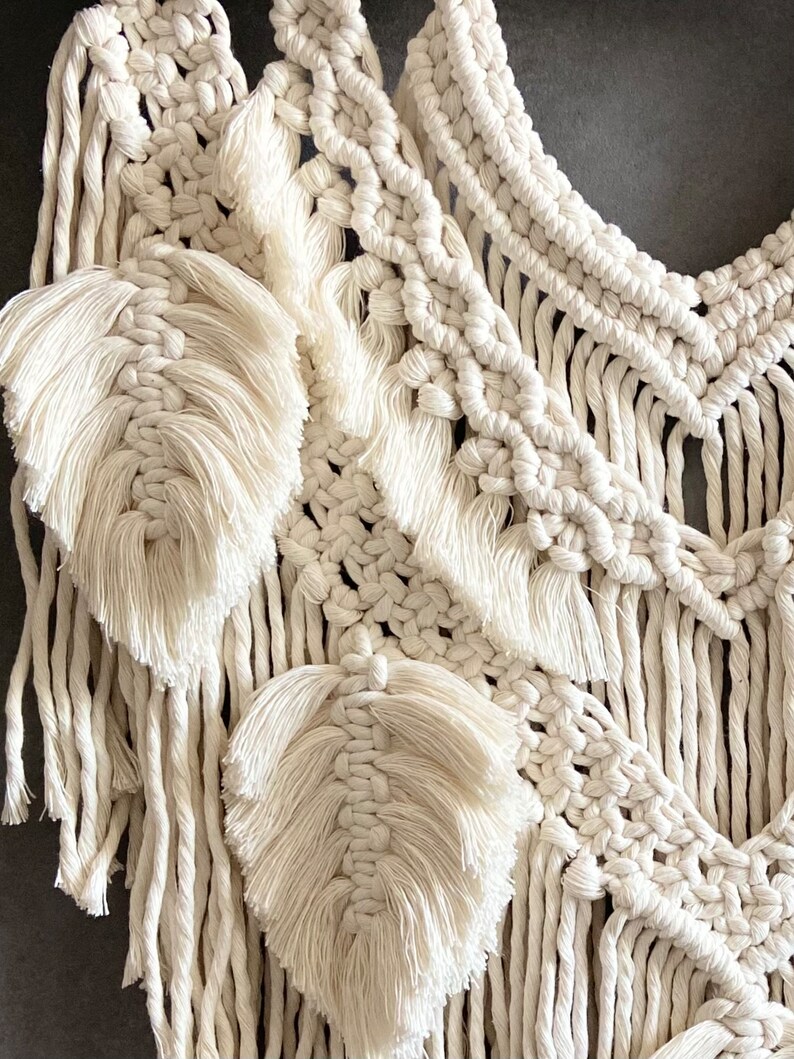 Puede incluir: Un tapiz de macram&eacute; blanco con un dise&ntilde;o geom&eacute;trico y detalles de flecos. El tapiz presenta una serie de hojas tejidas.