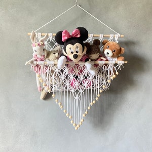 以下が含まれることがあります： 木製の棒とビーズが付いた白いマクラメの壁掛け。ハンガーには、ピンクと黒のミニーマウス、茶色のクマ、白いウサギなど、ぬいぐるみが詰められています。