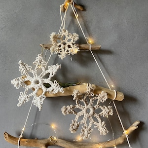 Puede incluir: Una decoración de árbol de Navidad de copo de nieve de macramé blanco con tres copos de nieve colgando de ramas de madera a la deriva con luces de hadas blancas.