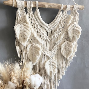 Puede incluir: Un tapiz de macram&eacute; beige con una barra de madera en la parte superior y un dise&ntilde;o de flecos. El tapiz presenta un patr&oacute;n geom&eacute;trico con formas de hojas.