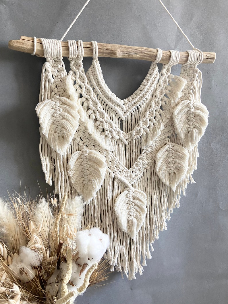 Puede incluir: Un tapiz de macram&eacute; beige con una barra de madera en la parte superior y un dise&ntilde;o de flecos. El tapiz presenta un patr&oacute;n geom&eacute;trico con formas de hojas.