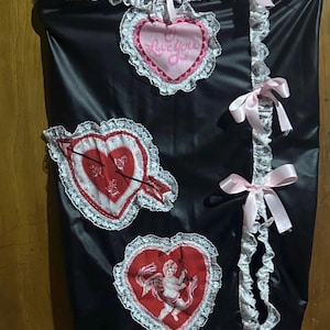 May include: Black skirt with white lace trim and pink bows. Features heart-shaped patches with designs: a heart pierced by an arrow, a heart with the text "Love You", and a heart with a cherub illustration.