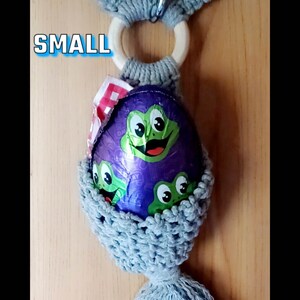 Op de afbeelding: Een grijze gehaakte paaseierenhouder met een houten ring aan de bovenkant en een lange kwast. De houder is gevuld met een paars chocolade-ei met groene kikkers erop. De tekst "SMALL" is zichtbaar in de afbeelding.