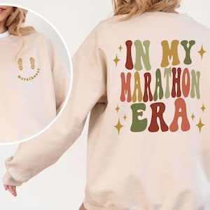 May include: A sand-colored crewneck sweatshirt featuring the text IN MY MARATHON ERA in large, wavy, multi-colored letters on the back, accented by gold stars. The front chest area displays a small graphic of two footprints and the word marathoner.